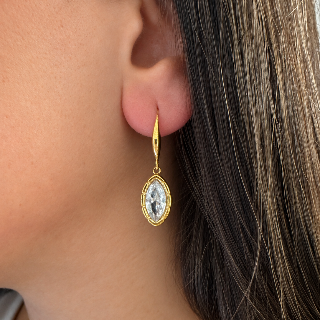 A close-up of a person's ear wearing a gold drop earring with a marquise-shaped clear gemstone in a gold bezel setting.