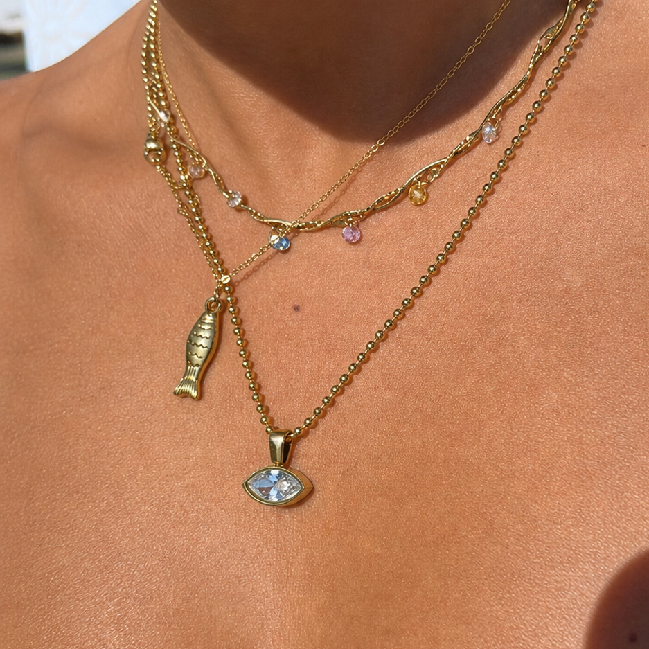 A close-up of a person wearing layered gold necklaces, including a fish-shaped pendant and a marquise-cut stone pendant, on a sunlit chest.